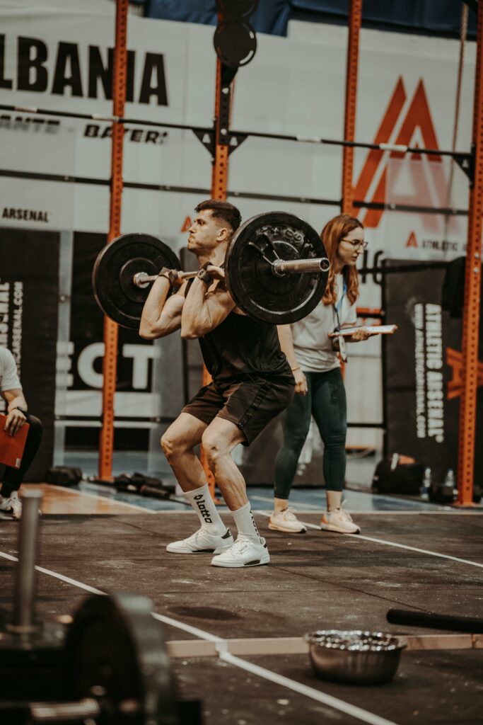 pexels-photo-16966372-16966372 Athlete lifting barbell in indoor gym competition setting with coach nearby.